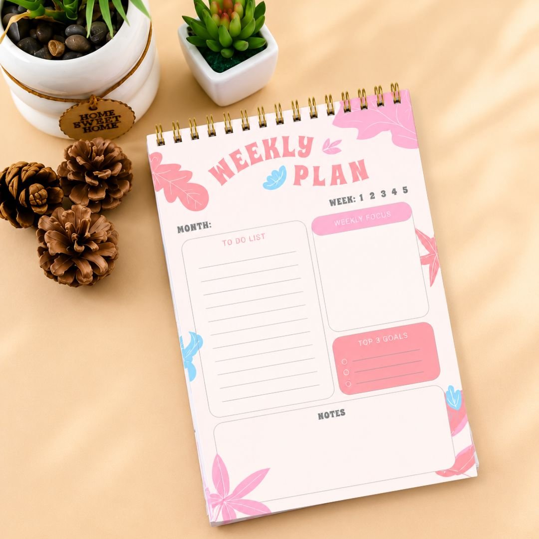 Weekly planner notepad pink spiral bound lifestyle on beige surface with succulents and pine cones